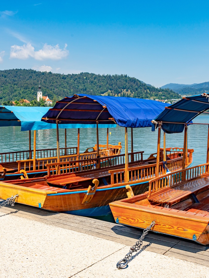 Holzboote mit blauen Schirmen, die an einem ruhigen See angedockt sind, mit Hügeln und Bäumen im Hintergrund.
