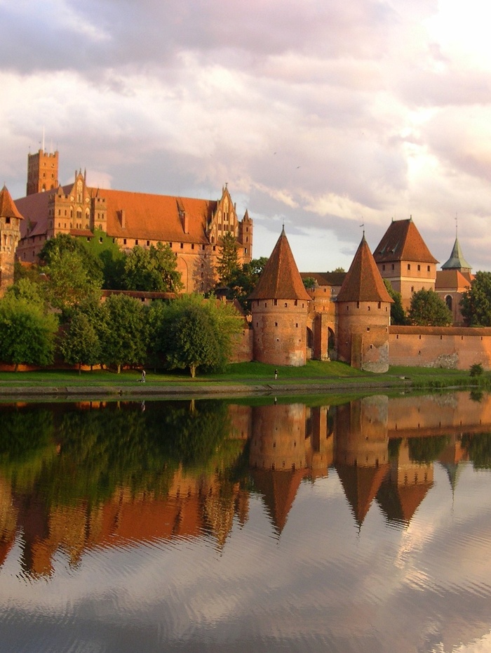 Eine Backsteinburg mit Türmen spiegelt sich in einem ruhigen Fluss unter einem bewölkten Himmel bei Sonnenuntergang.