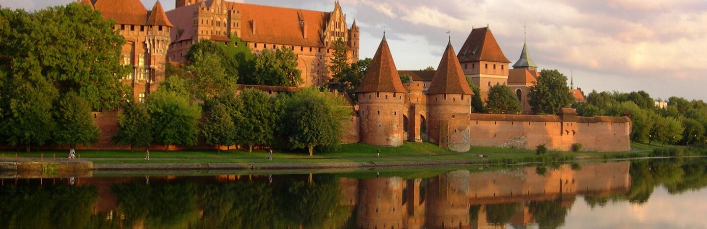 Eine Backsteinburg mit Türmen spiegelt sich in einem ruhigen Fluss unter einem bewölkten Himmel bei Sonnenuntergang.