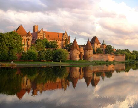 Eine Backsteinburg mit Türmen spiegelt sich in einem ruhigen Fluss unter einem bewölkten Himmel bei Sonnenuntergang.