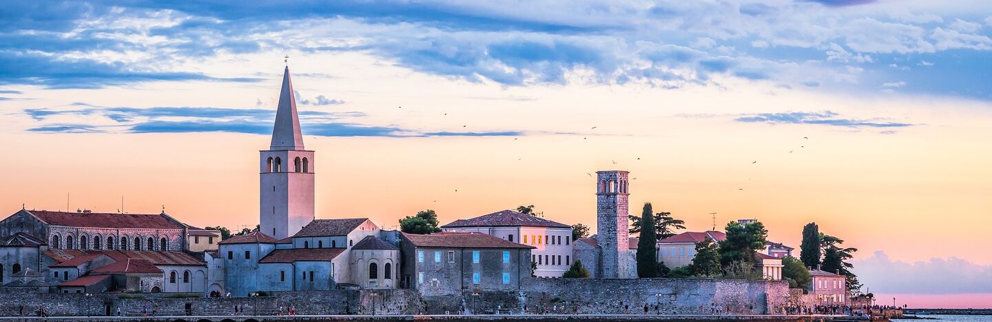 Küstenstadt bei Sonnenuntergang mit Kirchtürmen, historischen Gebäuden und ruhigem Wasser im Vordergrund.