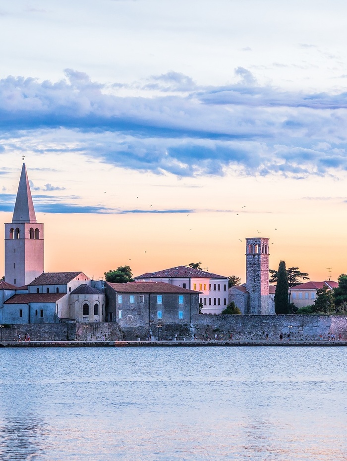 Küstenstadt bei Sonnenuntergang mit Kirchtürmen, historischen Gebäuden und ruhigem Wasser im Vordergrund.