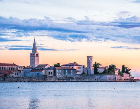 Küstenstadt bei Sonnenuntergang mit Kirchtürmen, historischen Gebäuden und ruhigem Wasser im Vordergrund.