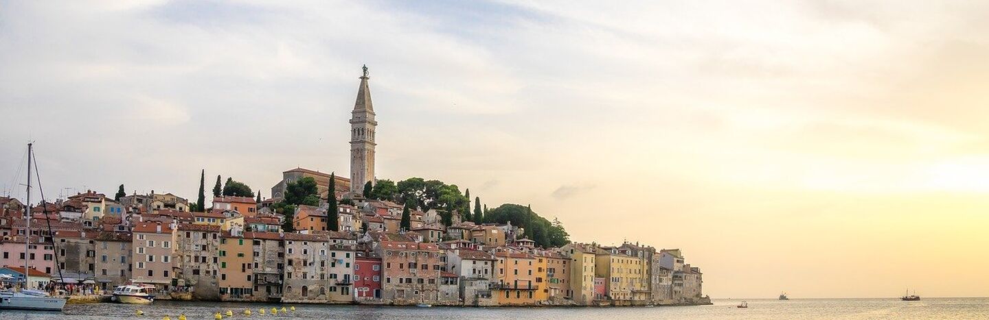 Ein weißes Kloster am See mit einem hohen Zwiebelturm vor dem Hintergrund von bewaldeten Bergen und blauem Himmel.