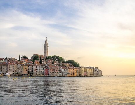 Ein weißes Kloster am See mit einem hohen Zwiebelturm vor dem Hintergrund von bewaldeten Bergen und blauem Himmel.