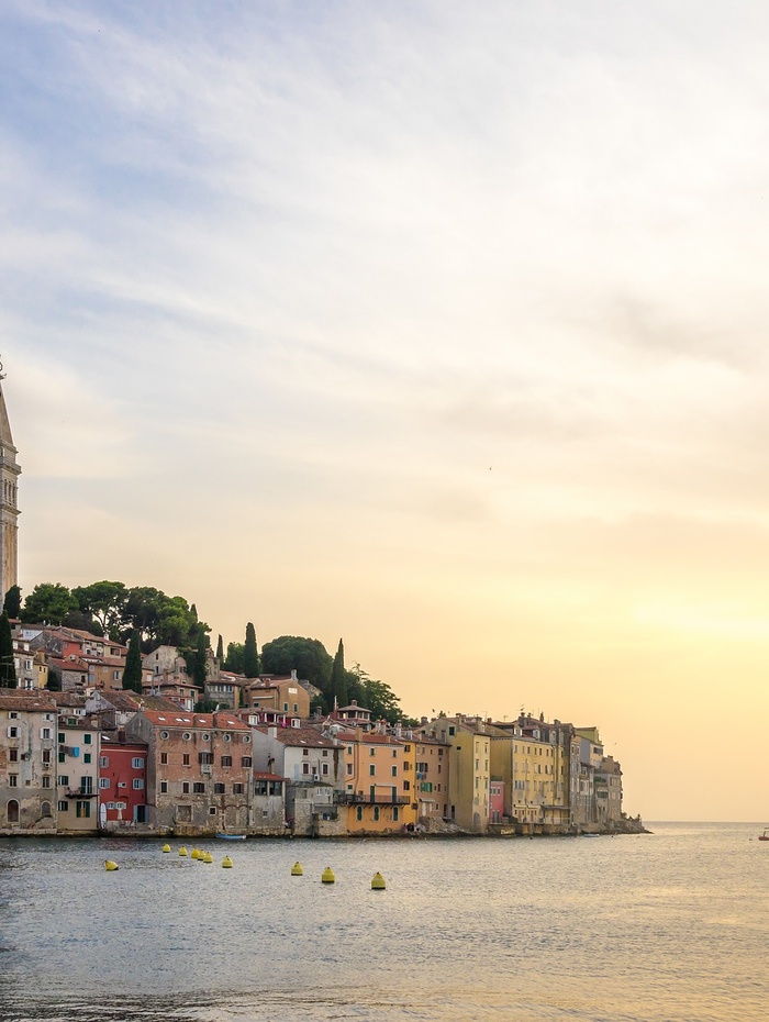 Bunte Gebäude am Meer und ein hoher Kirchturm bei Sonnenuntergang, mit Booten auf ruhigem Wasser im Vordergrund.