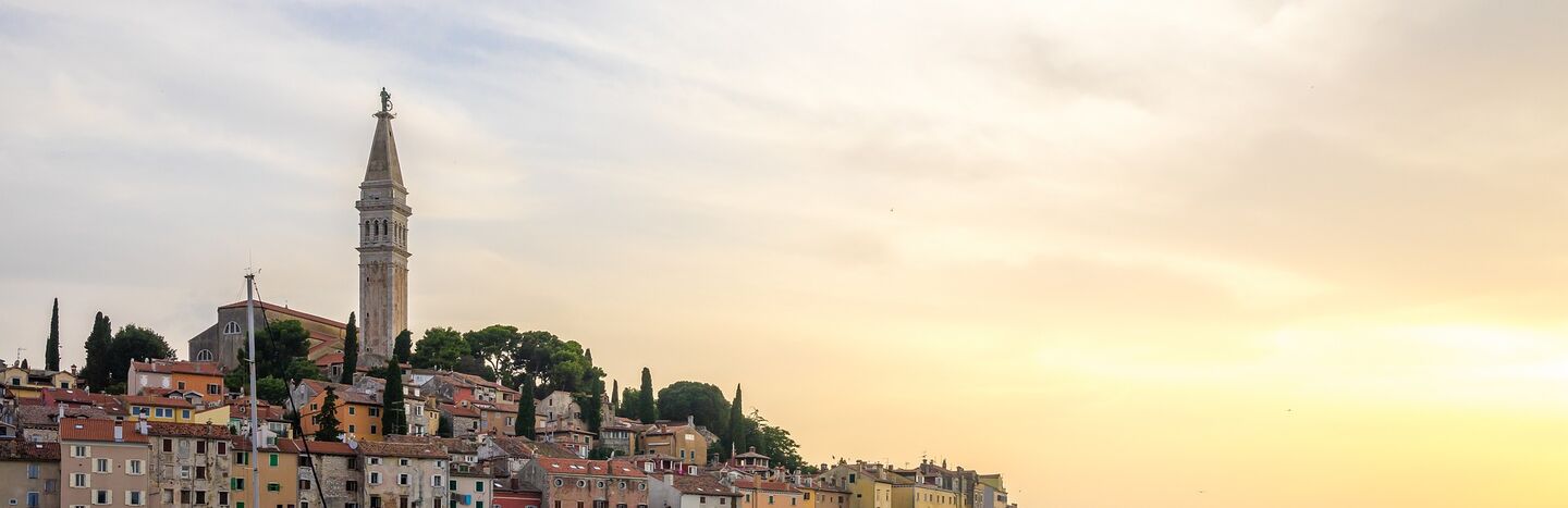 Bunte Gebäude am Meer und ein hoher Kirchturm bei Sonnenuntergang, mit Booten auf ruhigem Wasser im Vordergrund.