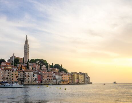 Bunte Gebäude am Meer und ein hoher Kirchturm bei Sonnenuntergang, mit Booten auf ruhigem Wasser im Vordergrund.