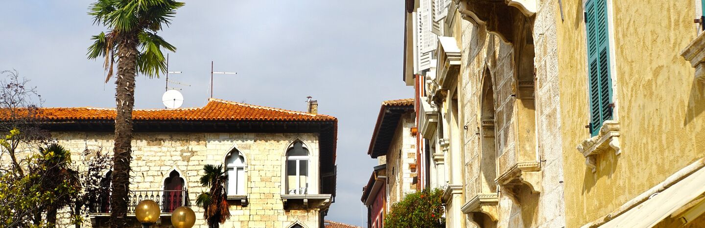 Alte europäische Gebäude mit gewölbten Fenstern und Palmen säumen eine sonnige Straße unter einem teilweise bewölkten Himmel.