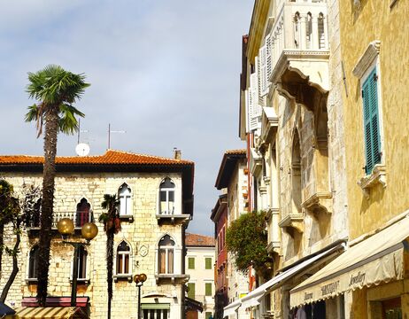 Alte europäische Gebäude mit gewölbten Fenstern und Palmen säumen eine sonnige Straße unter einem teilweise bewölkten Himmel.