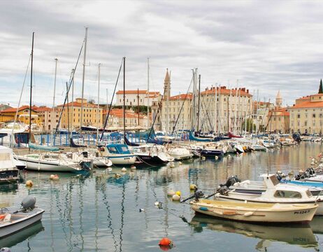 Segelboote und Yachten in einem Jachthafen mit Gebäuden und bewölktem Himmel im Hintergrund.