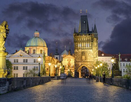 Die Karlsbrücke in Prag in der Abenddämmerung, mit historischen Gebäuden und Straßenlaternen, die bei bewölktem Himmel beleuchtet sind.