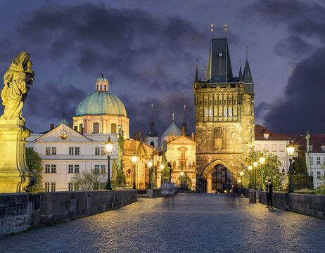 Die Karlsbrücke in Prag in der Abenddämmerung, mit historischen Gebäuden und Straßenlaternen, die bei bewölktem Himmel beleuchtet sind.