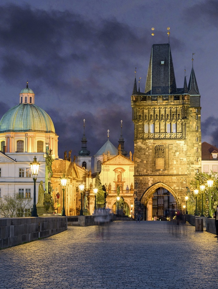 Die Karlsbrücke in Prag in der Abenddämmerung, mit historischen Gebäuden und Straßenlaternen, die bei bewölktem Himmel beleuchtet sind.