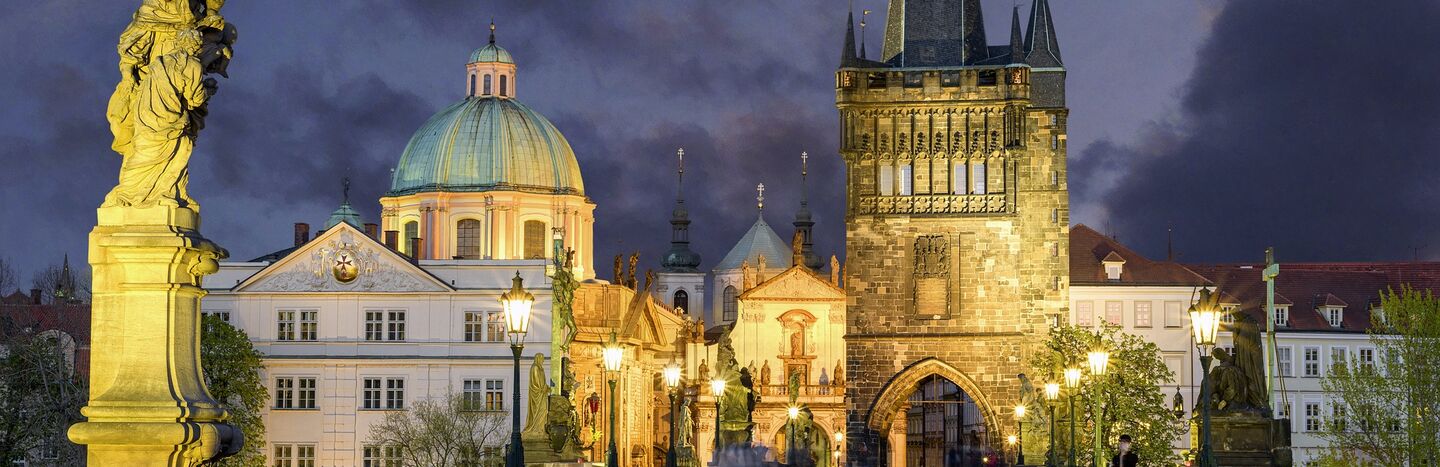 Die Karlsbrücke in Prag in der Abenddämmerung, mit historischen Gebäuden und Straßenlaternen, die bei bewölktem Himmel beleuchtet sind.