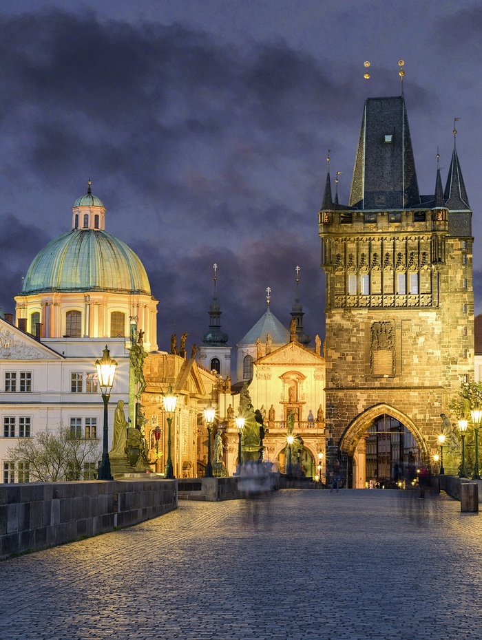Die Karlsbrücke in Prag in der Abenddämmerung, mit historischen Gebäuden und Straßenlaternen, die bei bewölktem Himmel beleuchtet sind.