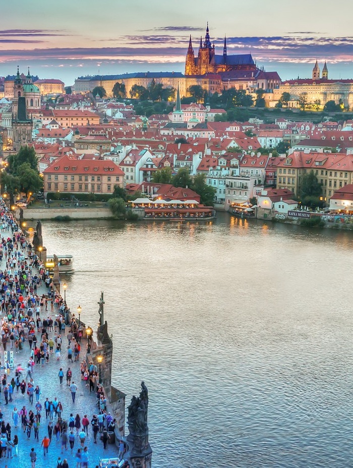 Menschenmassen laufen bei Sonnenuntergang über eine Brücke über einen Fluss mit einer Stadtlandschaft und einem historischen Schloss im Hintergrund.