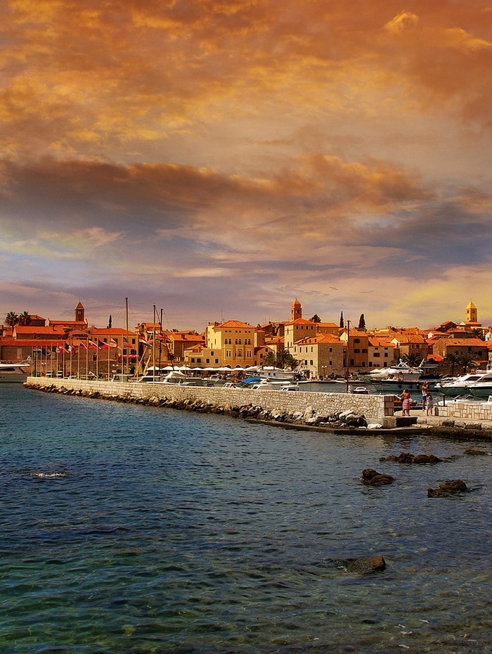 Ein farbenfroher Sonnenuntergang über einem Hafen mit Booten, steinernen Gebäuden und einem ruhigen, klaren blauen Meer.