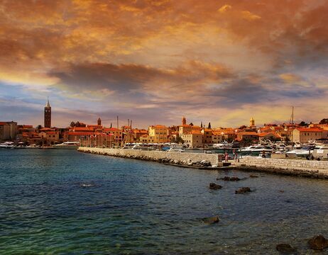 Ein farbenfroher Sonnenuntergang über einem Hafen mit Booten, steinernen Gebäuden und einem ruhigen, klaren blauen Meer.