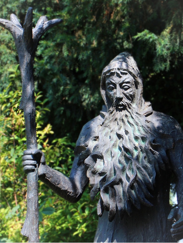 Bronzestatue eines bärtigen Mannes mit Kapuze, der einen hohen Stab hält, mit Bäumen im Hintergrund.