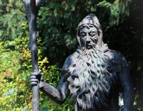 Bronzestatue eines bärtigen Mannes mit Kapuze, der einen hohen Stab hält, mit Bäumen im Hintergrund.