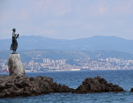 Statue einer Frau an einem felsigen Ufer mit einer Stadt und Bergen im Hintergrund.