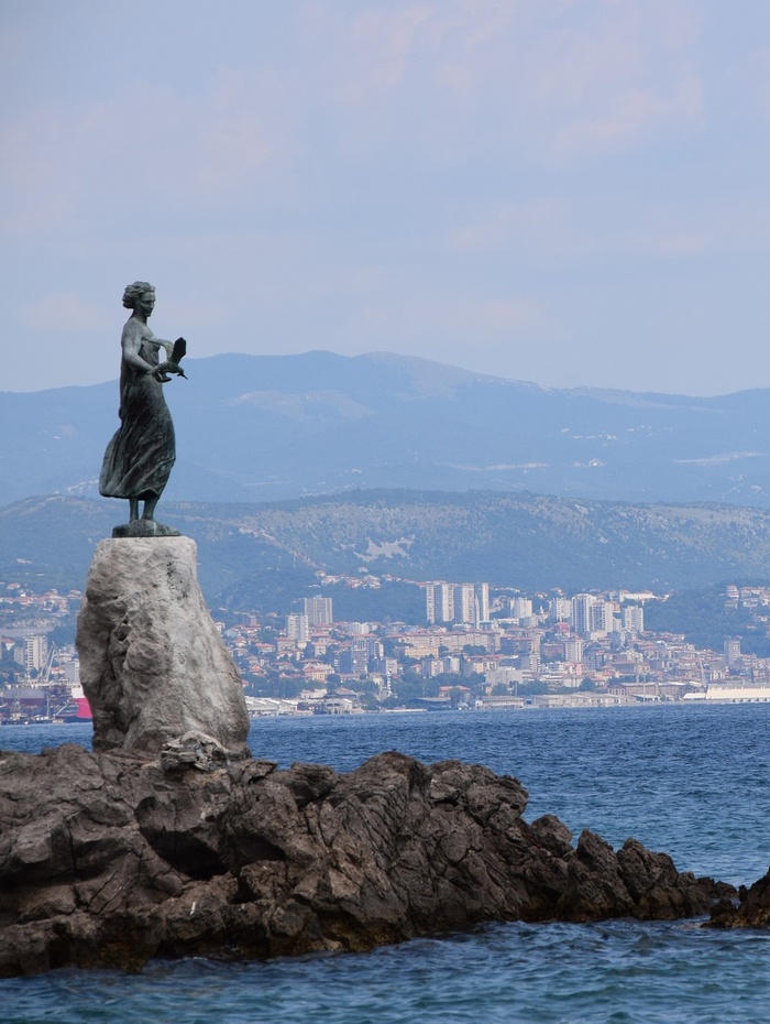 Statue einer Frau an einem felsigen Ufer mit einer Stadt und Bergen im Hintergrund.