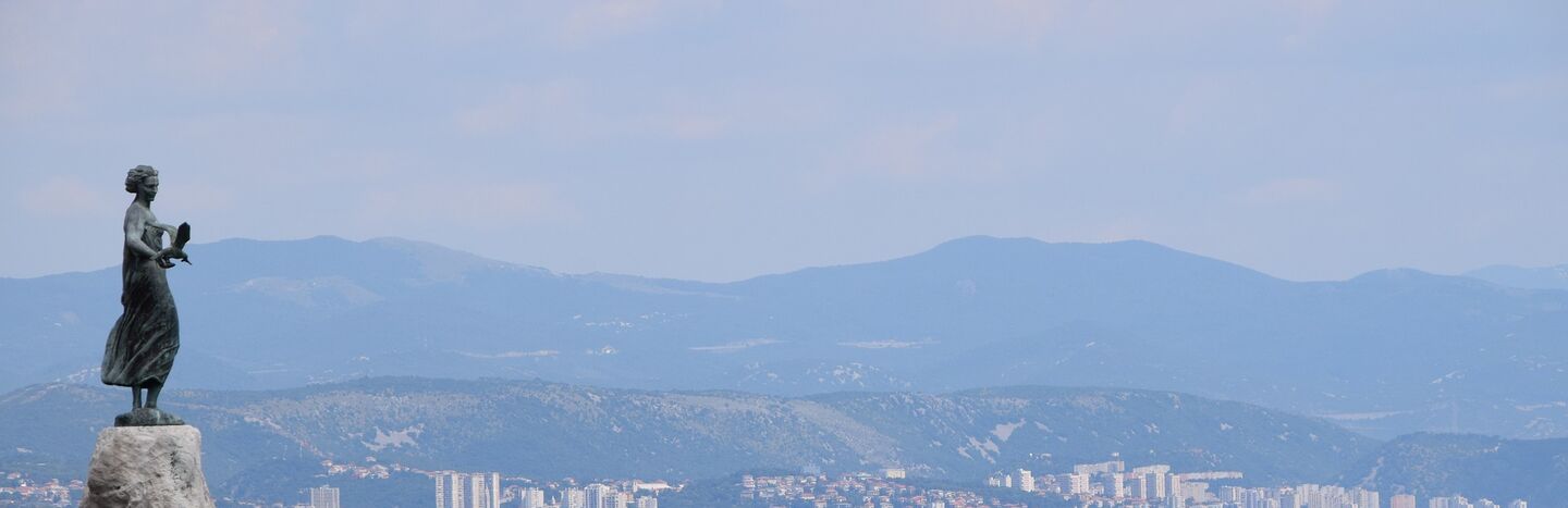 Statue einer Frau an einem felsigen Ufer mit einer Stadt und Bergen im Hintergrund.