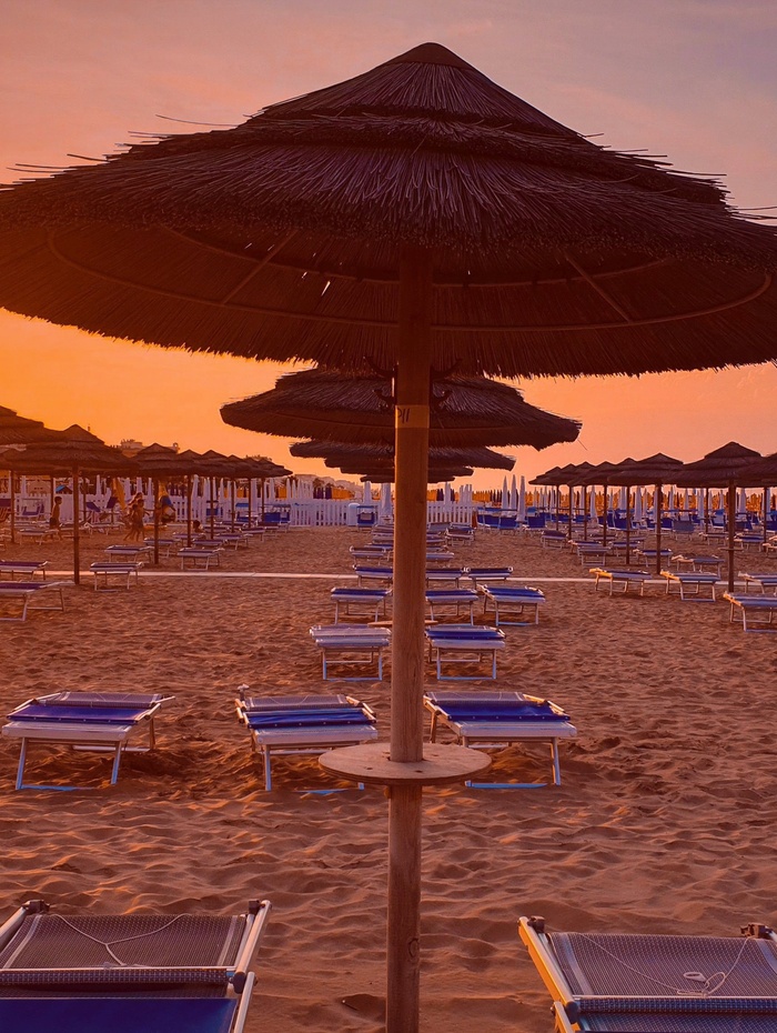 Reihen von Sonnenschirmen und Liegestühlen am Sandstrand bei Sonnenuntergang, mit warmer oranger Beleuchtung.