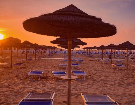 Reihen von Sonnenschirmen und Liegestühlen am Sandstrand bei Sonnenuntergang, mit warmer oranger Beleuchtung.