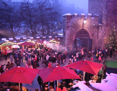 Eine Menschenmenge versammelt sich auf einem festlichen Markt im Freien in der Nähe einer Steinburg, mit roten Regenschirmen und Schnee auf dem Boden.