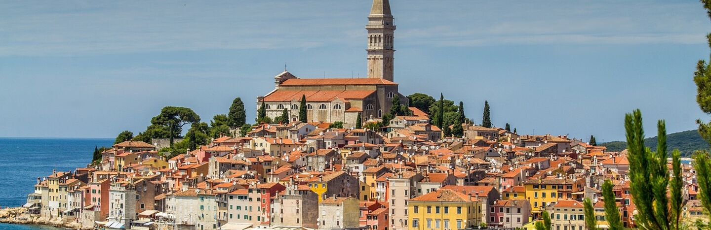 Buntes Küstenstädtchen mit einem hohen Kirchturm, Booten im Hafen und blauem Himmel darüber.