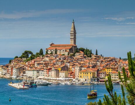 Buntes Küstenstädtchen mit einem hohen Kirchturm, Booten im Hafen und blauem Himmel darüber.