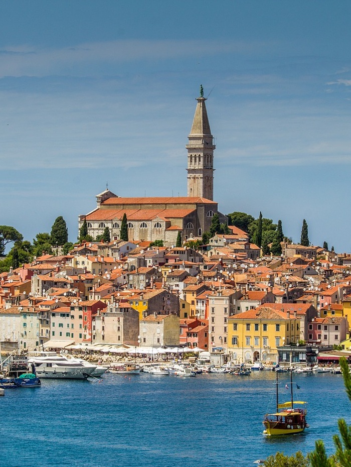 Buntes Küstenstädtchen mit einem hohen Kirchturm, Booten im Hafen und blauem Himmel darüber.