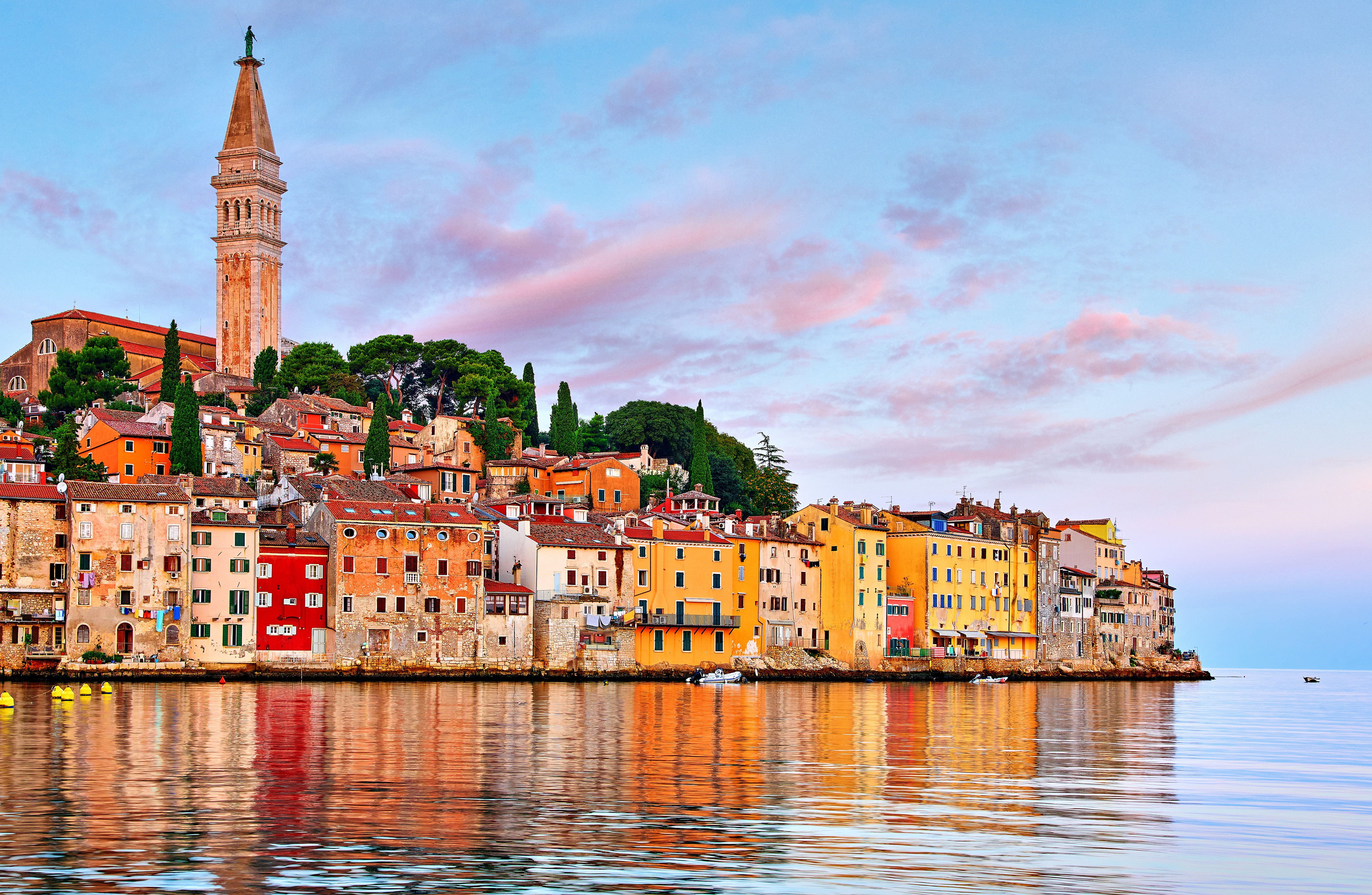 Bunte Gebäude am Meer und ein hoher Kirchturm spiegeln sich bei Sonnenuntergang auf dem ruhigen Wasser in Rovinj, Kroatien.
