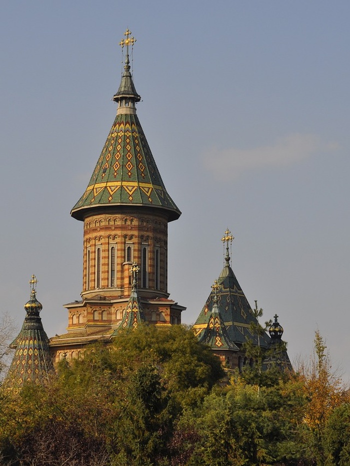 Verschnörkelte Kirche mit bunten Ziegeldächern und hohen Türmen, die teilweise von grünen Bäumen unter klarem Himmel verdeckt wird.