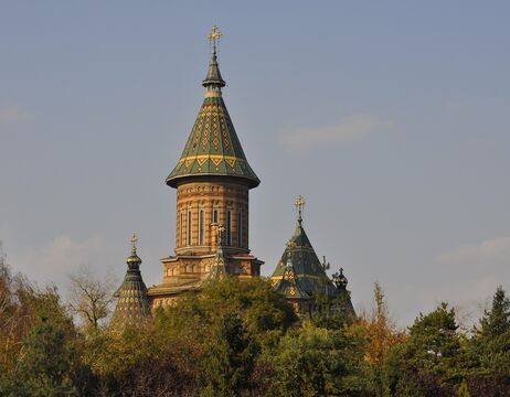 Verschnörkelte Kirche mit bunten Ziegeldächern und hohen Türmen, die teilweise von grünen Bäumen unter klarem Himmel verdeckt wird.
