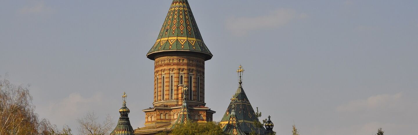 Verschnörkelte Kirche mit bunten Ziegeldächern und hohen Türmen, die teilweise von grünen Bäumen unter klarem Himmel verdeckt wird.