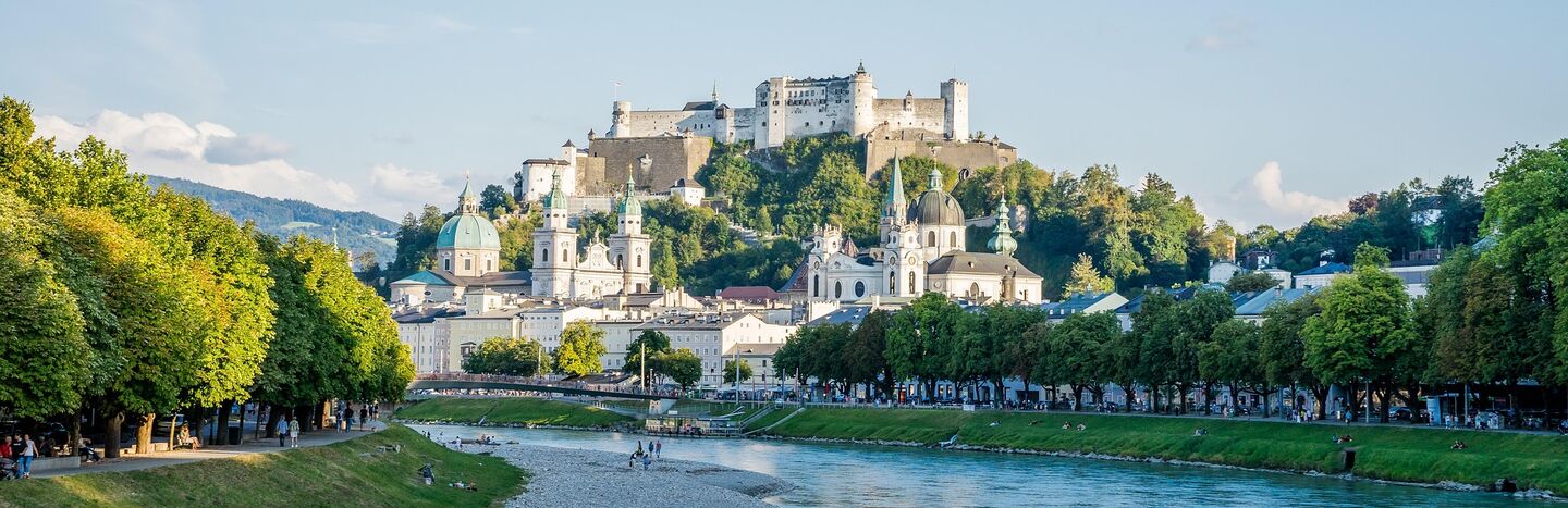 Ein Schloss auf einem Hügel mit Blick auf einen Fluss und historische Gebäude in einer europäischen Stadt an einem sonnigen Tag.