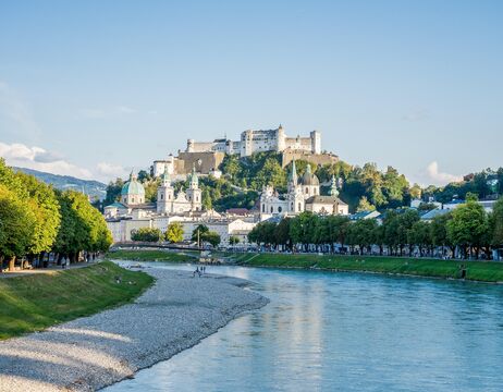 Ein Schloss auf einem Hügel mit Blick auf einen Fluss und historische Gebäude in einer europäischen Stadt an einem sonnigen Tag.