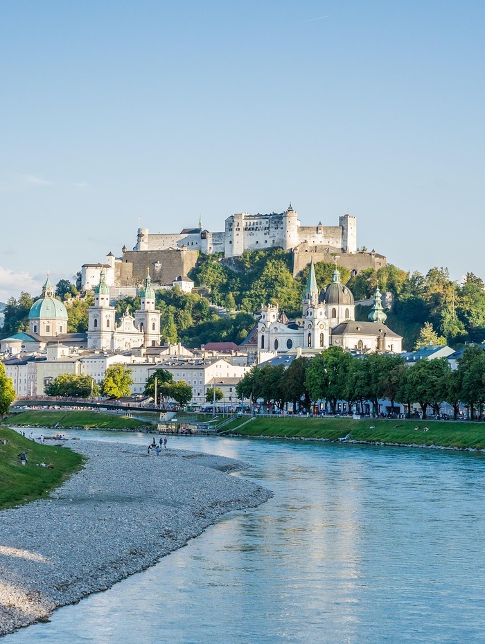 Ein Schloss auf einem Hügel mit Blick auf einen Fluss und historische Gebäude in einer europäischen Stadt an einem sonnigen Tag.