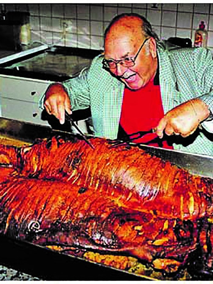 Ein lächelnder Mann mit Brille tranchiert ein großes gebratenes Schwein auf einer Küchenzeile.
