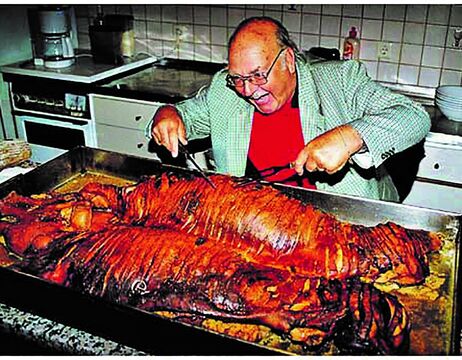 Ein lächelnder Mann mit Brille tranchiert ein großes gebratenes Schwein auf einer Küchenzeile.