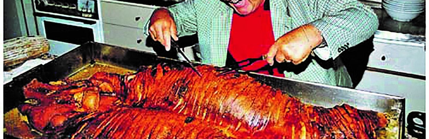 Ein lächelnder Mann mit Brille tranchiert ein großes gebratenes Schwein auf einer Küchenzeile.