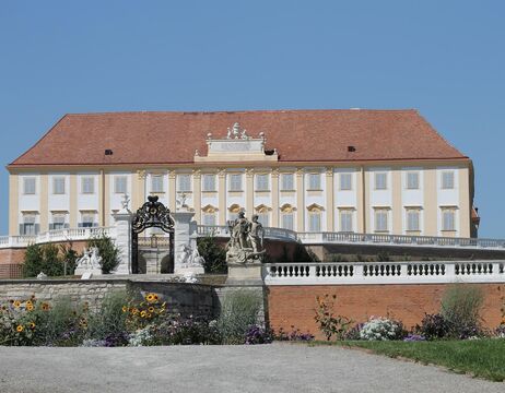 Ein Gebäude im Barockstil mit einer Statue, einem verzierten Tor und bunten Blumen im Vordergrund an einem sonnigen Tag.