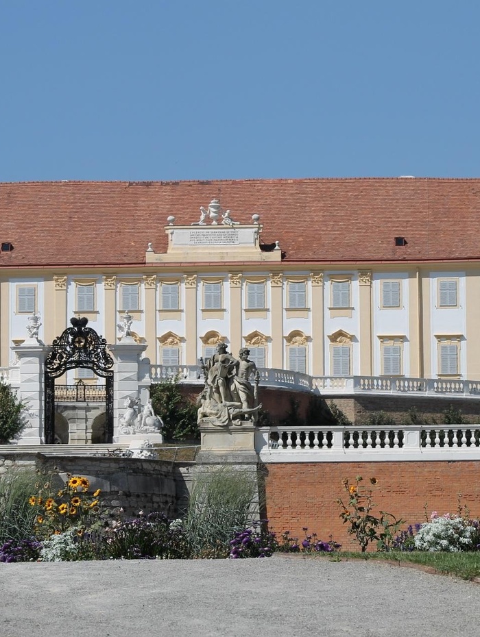 Ein Gebäude im Barockstil mit einer Statue, einem verzierten Tor und bunten Blumen im Vordergrund an einem sonnigen Tag.