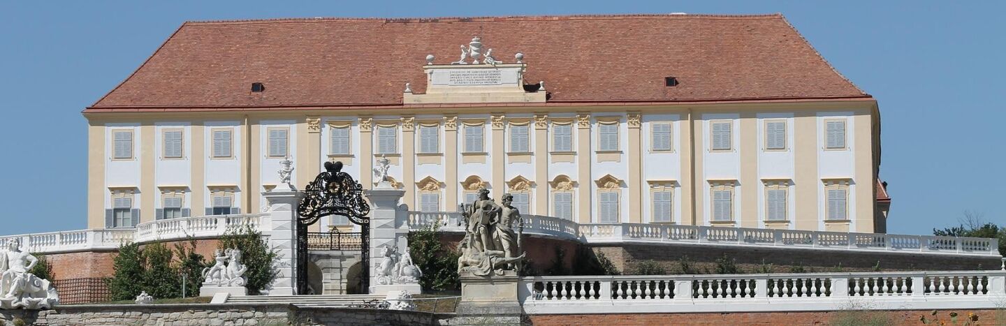 Ein Gebäude im Barockstil mit einer Statue, einem verzierten Tor und bunten Blumen im Vordergrund an einem sonnigen Tag.