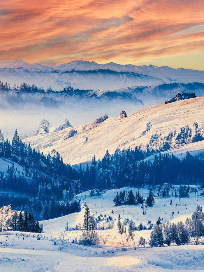 Schneebedeckte Hügel und Kiefern mit fernen Bergen unter einem farbenfrohen Sonnenuntergangshimmel.