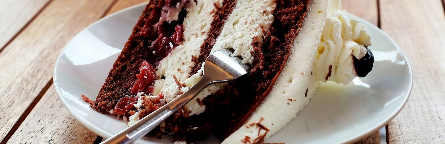 Eine Gabel liegt auf einem Teller, auf dem ein Stück Schokoladen-Sahne-Torte mit Kirschen liegt.