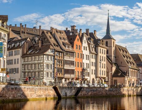 Bunte historische Gebäude und eine Kirche säumen einen Kanal unter einem teilweise bewölkten Himmel in einer europäischen Stadt.